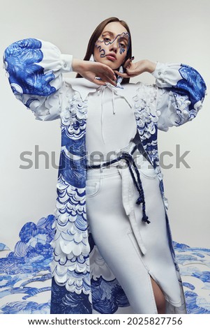 Woman in amazing dress with blue and white waves and wire mask. Drawing and ruffles on clothes. Model standing on the blue waves drawing                                              
