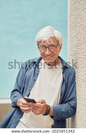 Vertical portrait of modern senior man using smartphone outdoors and smiling at camera while standing against light blue background