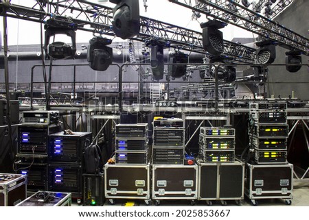 Installation of professional sound, light and stage equipment for a concert. Backstage and tech zone with rack amplifiers, audio signal splitters, radio microphone systems and flight cases. Royalty-Free Stock Photo #2025853667