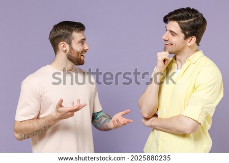 Two pensive young men 20s friends together wearing casual t-shirt tattoo translate fun talking speak prop up chin spread hands arms look aside isolated on purple background. People lifestyle concept. Royalty-Free Stock Photo #2025880235