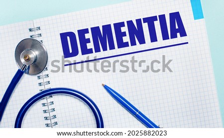 On a light blue background, an open notebook with the word DEMENTIA, a blue pen and a stethoscope. View from above. Medical concept