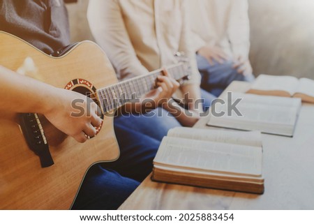 A young man is playing guitar and sings a song from a Christian hymn book with his friends at home, Christian family worship concept Royalty-Free Stock Photo #2025883454