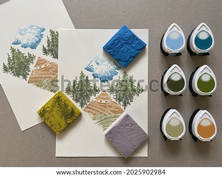 A colourful pattern of mountains, pines and a cottage printed on paper, with the hand-carved rubber stamps, and the green, blue and yellow colored ink pads used for the process. Royalty-Free Stock Photo #2025902984
