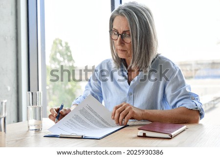Serious older lawyer businesswoman in glasses reading signing trust partnership contract sit at table in office. Executive ceo put signature make legal bank sale financial loan investment agreement. Royalty-Free Stock Photo #2025930386