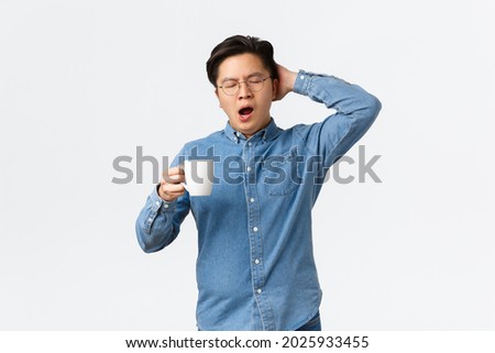 Portrait of sleepy young male employee yawning and holding mug with coffee, waking-up tired. Man working late wanting sleep, holding head and close eyes, standing white background Royalty-Free Stock Photo #2025933455