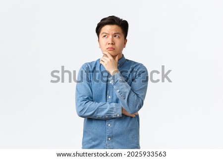 Thoughtful complicated asian man in shirt, touching chin and looking upper left corner, thinking, making decision, choosing something, having doubts, standing hesitant over white background Royalty-Free Stock Photo #2025933563