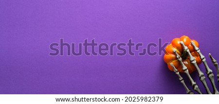 Happy Halloween holiday banner template. Flat lay skeletons hand with pumpkin on purple background. Top view with copy space.
