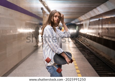 Young girl in casual shirt and jeans stand on platform wait for train arrival. Female student skateboarder at underground metro station travel home with longboard by public transport. Woman in subway