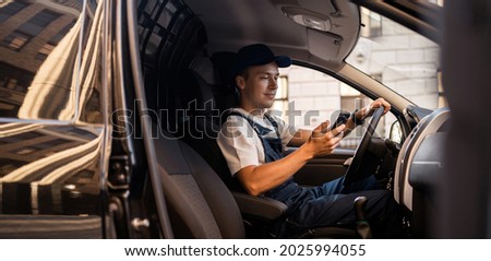 In the form of a courier, a man of Caucasian appearance works on the phone while driving a car. Transport company an experienced driver delivers the goods. The entrepreneur is young in his business. Royalty-Free Stock Photo #2025994055