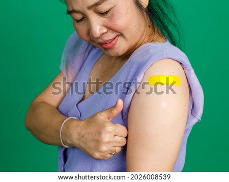 Studio close up shot of Asian female patient hand showing thumb up with yellow medical bandage plaster after receiving coronavirus covid 19 vaccination from doctor at hospital on green background.