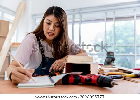 Portrait of young asian female carpenter assempling DIY wooden furniture and making notes on her notebook with workshop background. Concept female worker.
