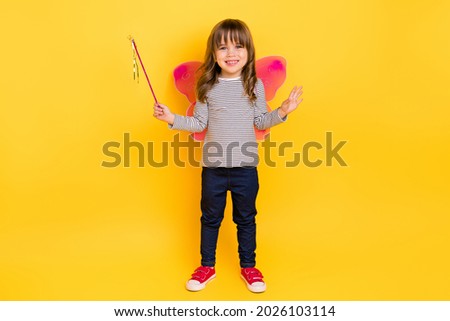 Full length photo of cute small blond hair girl hold stick wear wings shirt jeans sneakers isolated on yellow backgound