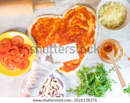 Hand in glove holding empty business card on hear shape pizza dough with tomato sauce background. Ingredients for cooking pizza with dough in heart shape