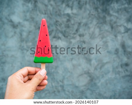 Hand holding artificial watermelon slice popsicle on grey concrete wall background with copy space. Summer fruit ice cream stick.