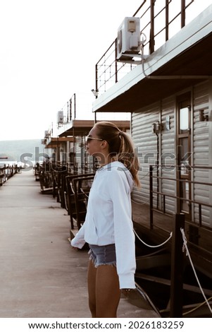 portrait of a beautiful blonde girl on the pier, dressed in white sweatshirt, short denim shorts and sunglasses, lifestyle model at the river side, summer beach vocation at the water