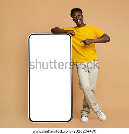 Happy Young Black Man Leaning And Pointing At Big Smartphone With White Blank Screen, Cheerful African Guy Showing Free Copy Space For Your Design, Standing Over Beige Background, Mockup Image Royalty-Free Stock Photo #2026204490