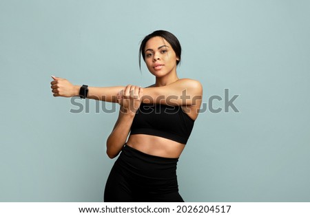 Warm up concept. Young slim african american woman stretching her arms before training, standing over blue background and looking at camera, wearing activity tracker Royalty-Free Stock Photo #2026204517