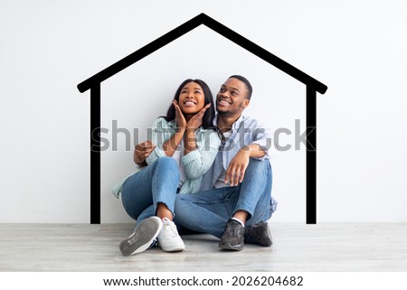 Happy african american spouses sitting near white wall with drawn house, dreaming and imagining their new home, planning relocation, creative collage Royalty-Free Stock Photo #2026204682