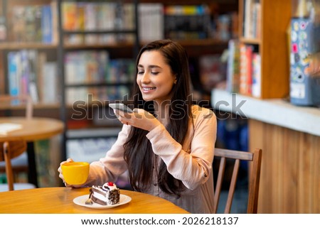 Excited arab lady taking photo of coffee and dessert while having breakfast at cafe. Lovely woman making photo of her meal, creating happy memories at restaurant