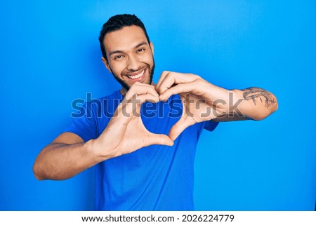 Hispanic man with beard wearing casual blue t shirt smiling in love doing heart symbol shape with hands. romantic concept. 