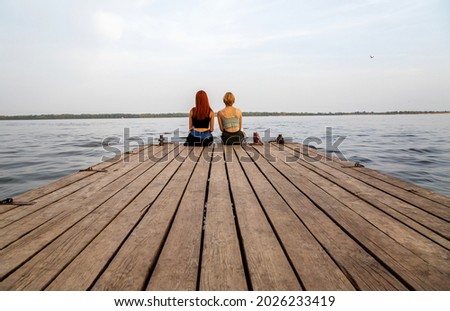 Two girls sitting on a wooden pier on the background of water. Vacation and travel, summer vacation and dreams.