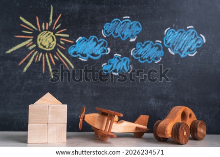 A wooden house and a toy airplane and a car stand against a chalk board with a picture of the sun and clouds. The concept of returning home from a trip