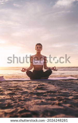 Woman sitting on exercise mat practicing yoga on the beach. Caucasian woman meditating in lotus pose.