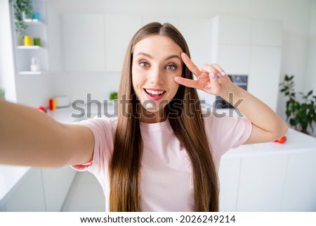 Photo portrait young woman smiling taking selfie in kitchen at home showing v-sign gesture