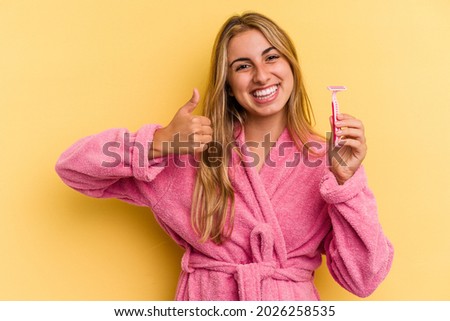 Young caucasian blonde woman wearing bathrobe holding blades isolated on yellow background  smiling and raising thumb up
