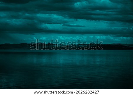   Blue green clouds over the sea. Toned seascape. Dark teal water and sky background with copy space for design.Rocky coastline on the horizon. Creepy, scary atmosphere, mood.                          Royalty-Free Stock Photo #2026282427