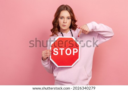 Portrait of displeased curly haired teenage girl in hoodie pointing finger at stop sign, strictly looking camera. Indoor studio shot isolated on pink background