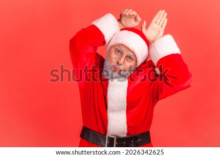 Funny elderly man with gray beard wearing santa claus costume pout lips playfully and showing bunny ears, aping with comical childish grimace. Indoor studio shot isolated on red background.