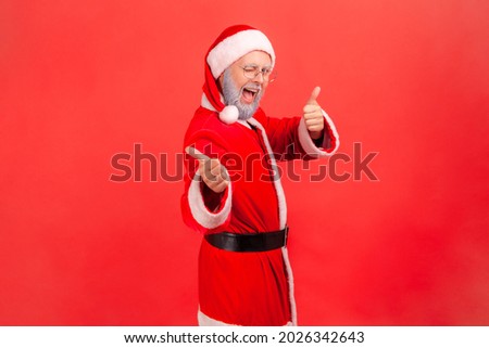 Profile portrait of elderly man with gray beard wearing santa claus costume looking at camera with excited look, winking and showing thumb up. Indoor studio shot isolated on red background.