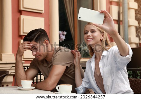 Young woman trying to take selfie with her displeased boyfriend in outdoor cafe. Boring date Royalty-Free Stock Photo #2026344257
