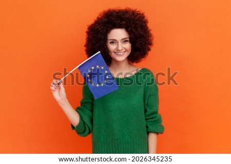 Smiling woman with Afro hairstyle wearing green casual style sweater holding Europe flag, symbol of Europe, EU association and community. Indoor studio shot isolated on orange background.