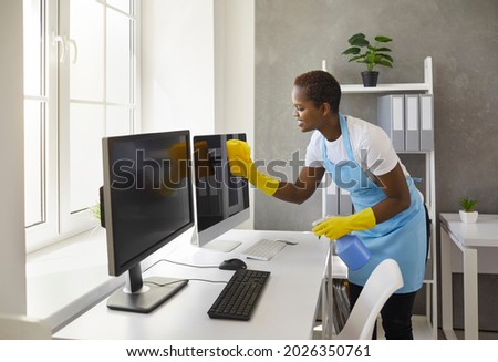 Smiley caretaker, charwoman, daily janitor service lady in uniform with antiseptic spray bottle and wet cloth rag cleaning desktop PC computer, desk and table surfaces in modern office room interior Royalty-Free Stock Photo #2026350761