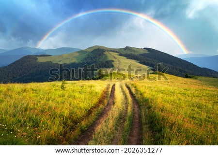 Amazing scene in summer mountains. Lush green grassy meadows in fantastic evening sunlight. Rural road and beautyful rainbow in dramatic sky. Landscape photography Royalty-Free Stock Photo #2026351277
