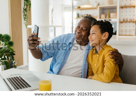 Happy African grandad and grandson sitting at home and taking a selfie with the new phone. They are so happy together that they must photograph it.