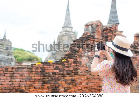 Rear view woman Asian tourist wearing a surgical mask taking a pictures in historical park with old ancient capital pagoda or stupa background. Travel during Covid-19 pandemic and new normal concept.