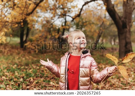Little blonde girl plays with yellow autumn leaves in the garden, smile, has fun