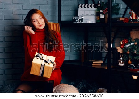 Asian girl with gift in her hands. Pretty woman in red knitted sweater. Christmas tree and decorations on shelf. New year mood. Gray brick wall in background.