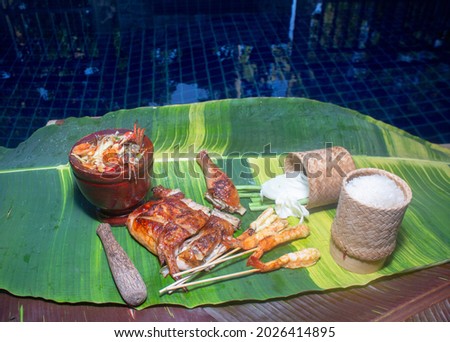 
Sticky rice, papaya salad, grilled chicken and grilled shrimp served on a spotted banana leaf poolside.
red yellow green speckled banana leaves in the background. food and restaurant concept.

