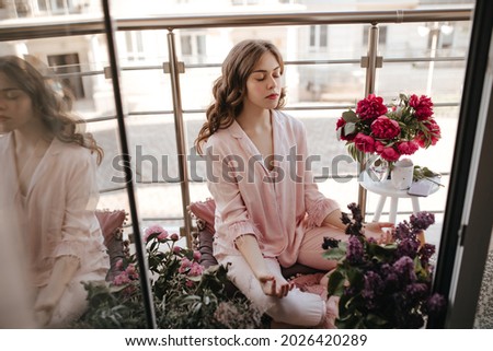 Young pleasent moldavian female meditating among fresh flowers with closed eyes. Brunette girl with natural face in wonderful pink chiffon homewear relaxing on her cozy balcony with beautiful sunlight