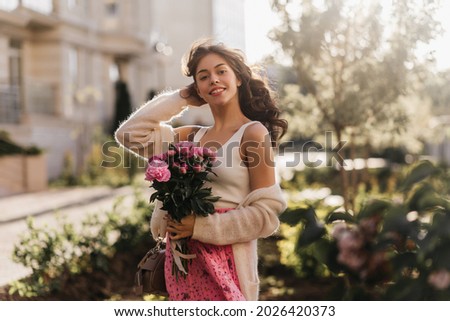 Medium portrait of adolescent female smiling and posing in front of the camera. Gorgerous sunlight highlights her rosy printed skirt and milky singlet, posing with a bouquet of peonies