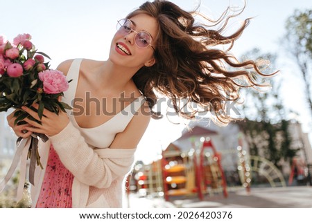 Image of young lady walking in the park with lovely flowers and sunny weather. Playing with her hair and smiling sincerely to the camera, wearing classy outfit and natural make up