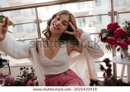 Portrait of young european lady showing peace sign and taking selfie. Sitting on the veranda floor next flower bouquets in white singlet and vintage milky cardigan with red lipstick and smiling widely