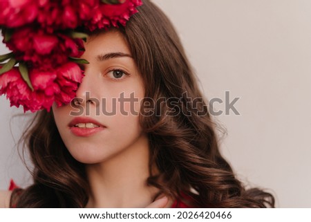 Full close up photography of brown hair grey eyes lady watching directly into the camera. Wonderfull peonies bouquet, summer cherry lipstick. High quality photo above white wall with soft light