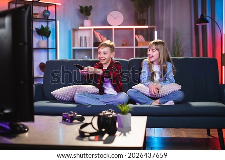 Pretty brother and sister sitting with crossed legs on comfy couch and watching TV. Happy two children in casual wear enjoying funny cartoons or interesting film at home. Royalty-Free Stock Photo #2026437659