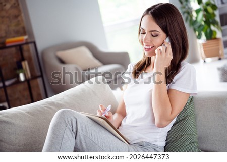 Photo portrait brunette woman sitting on couch at home smiling talking on mobile phone writing notes in notebook
