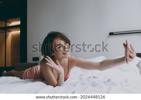 Full body young woman in pajamas lying in bed do selfie shot on mobile cell phone camera waving hand talk by video call rest relax on weekends indoors at home. Good mood night morning bedtime concept.